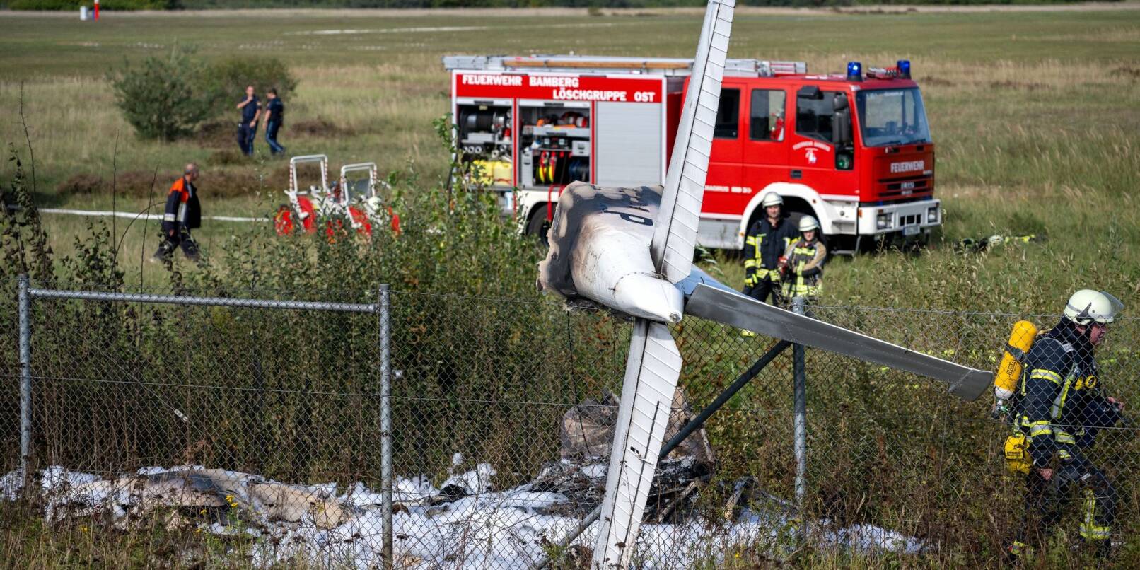 Trümmerteile eines Flugzeugs hängen in einem Zaun, während Einsatzkräfte der Feuerwehr an der Absturzstelle stehen.