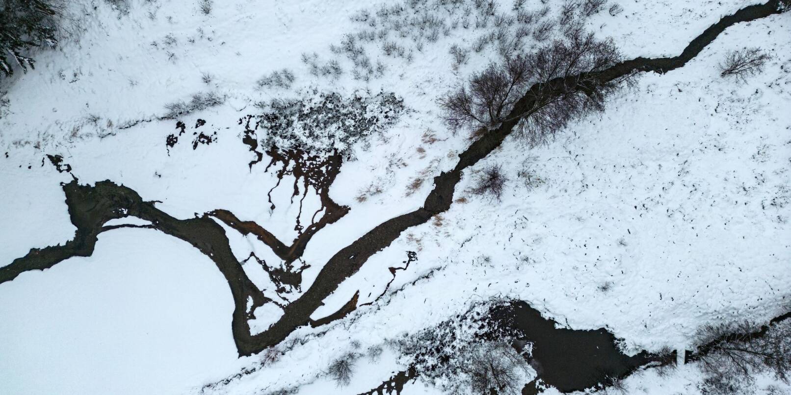 Ein Bachlauf schlängelt sich durch die verschneite Winterlandschaft im Taunus (Luftaufnahme mit einer Drohne).