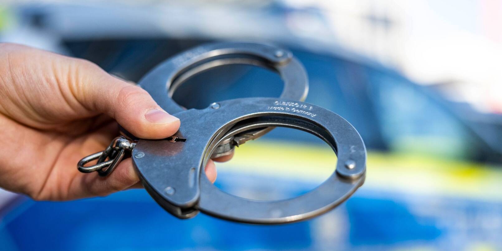 Eine Hand hält Handschellen vor einen Streifenwagen der Polizei. Eine Hand hält Handschellen vor einen Streifenwagen der Polizei.