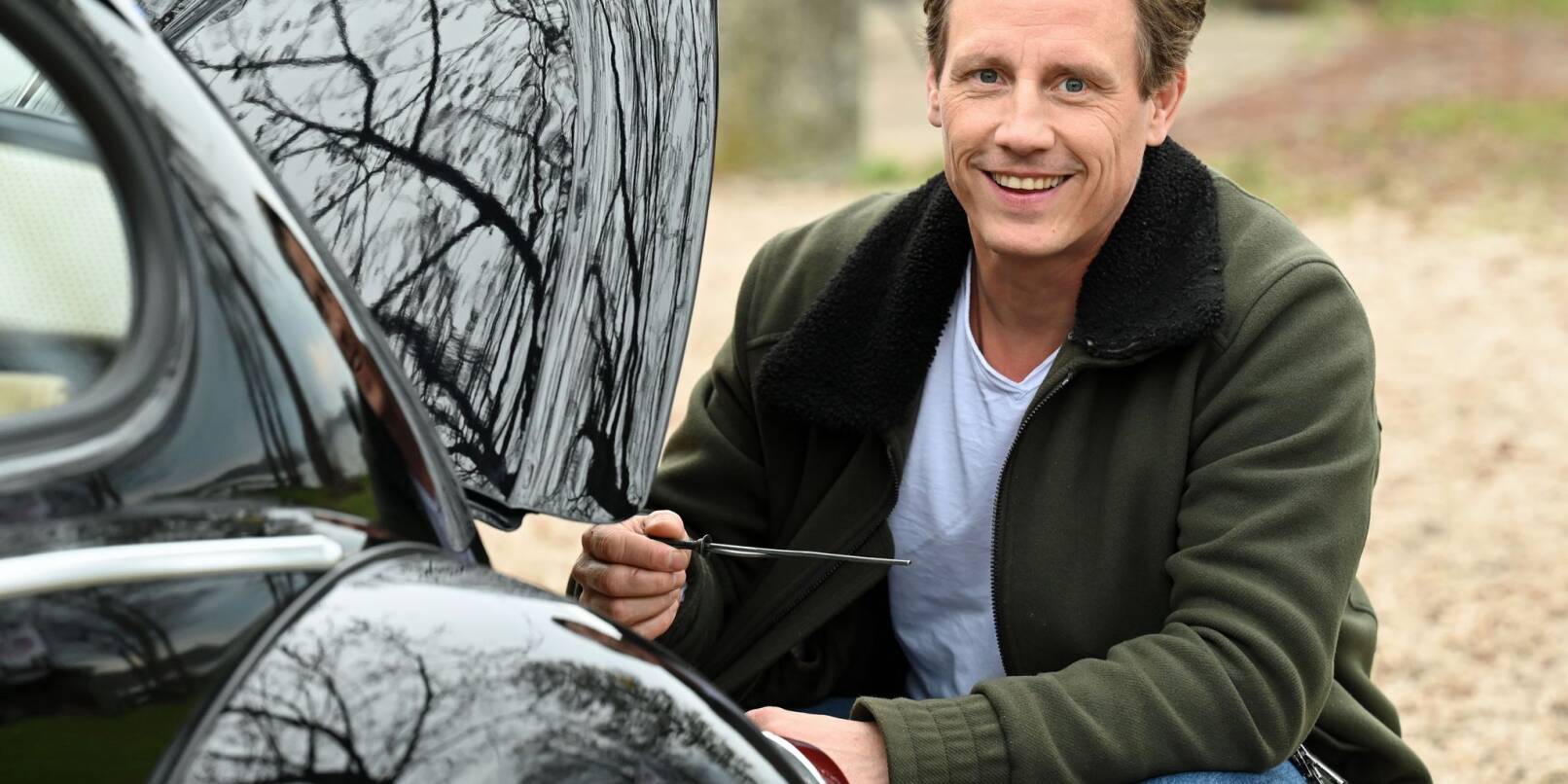 Der Schauspieler Patrick Kalupa hockt bei einem Fototermin an seinem Oldtimer VW Käfer.