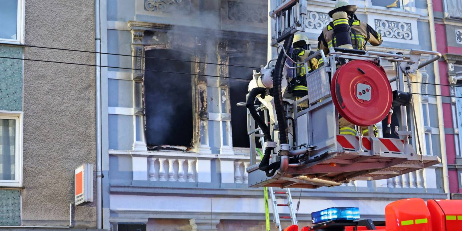 Feuerwehrmänner stehen vor dem Mehrfamilienhaus in Solingen; Rauch dringt aus einem der Fenster.