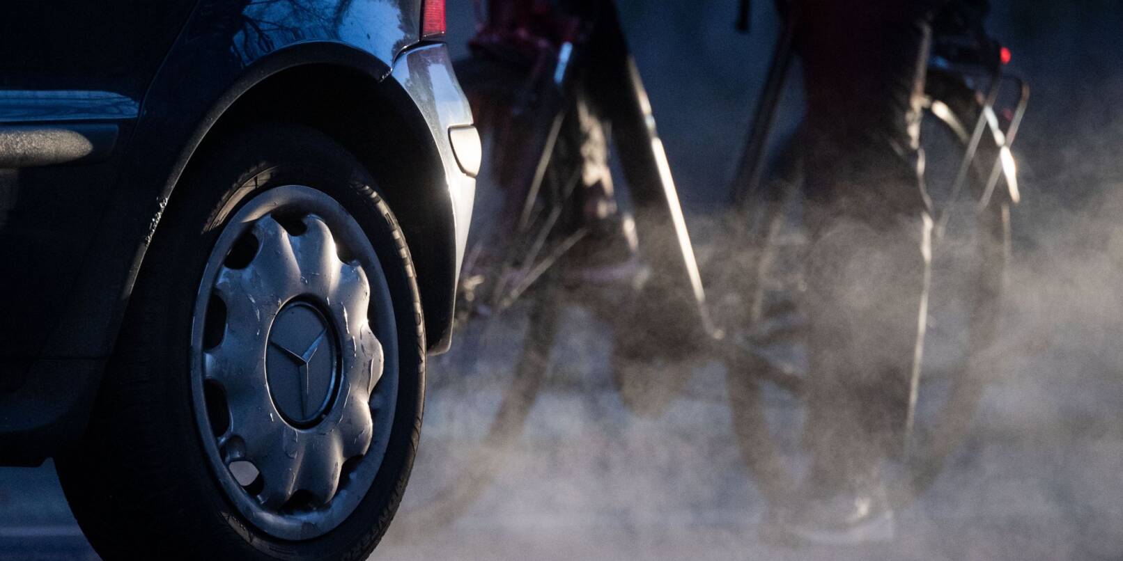 Ein Radfahrer steht neben einem Auto von Mercedes mit Dieselantrieb, dessen Abgase in der kalten Morgenluft sichtbar werden.