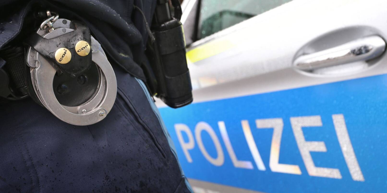 Ein Polizist mit Handschellen und einer Pistole am Gürtel steht vor einem Streifenwagen. Ein Polizist mit Handschellen und einer Pistole am Gürtel steht vor einem Streifenwagen.